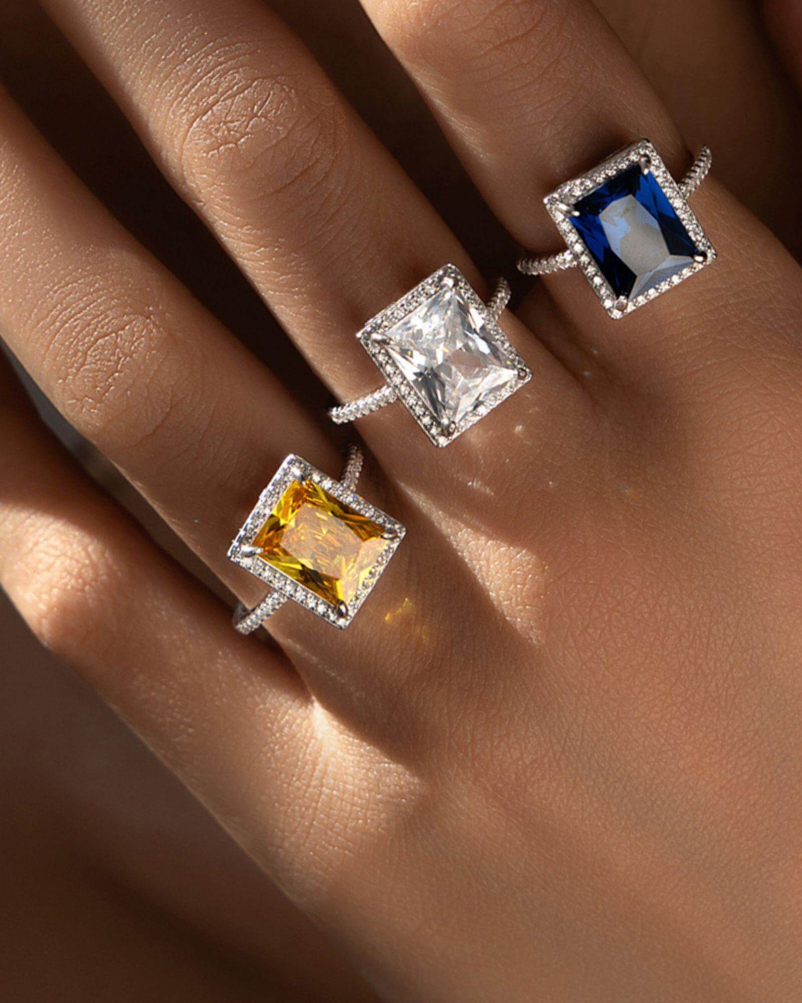 Three radiant cut cocktail rings in silver featuring blue, yellow, and clear zirconia gemstones on a hand.