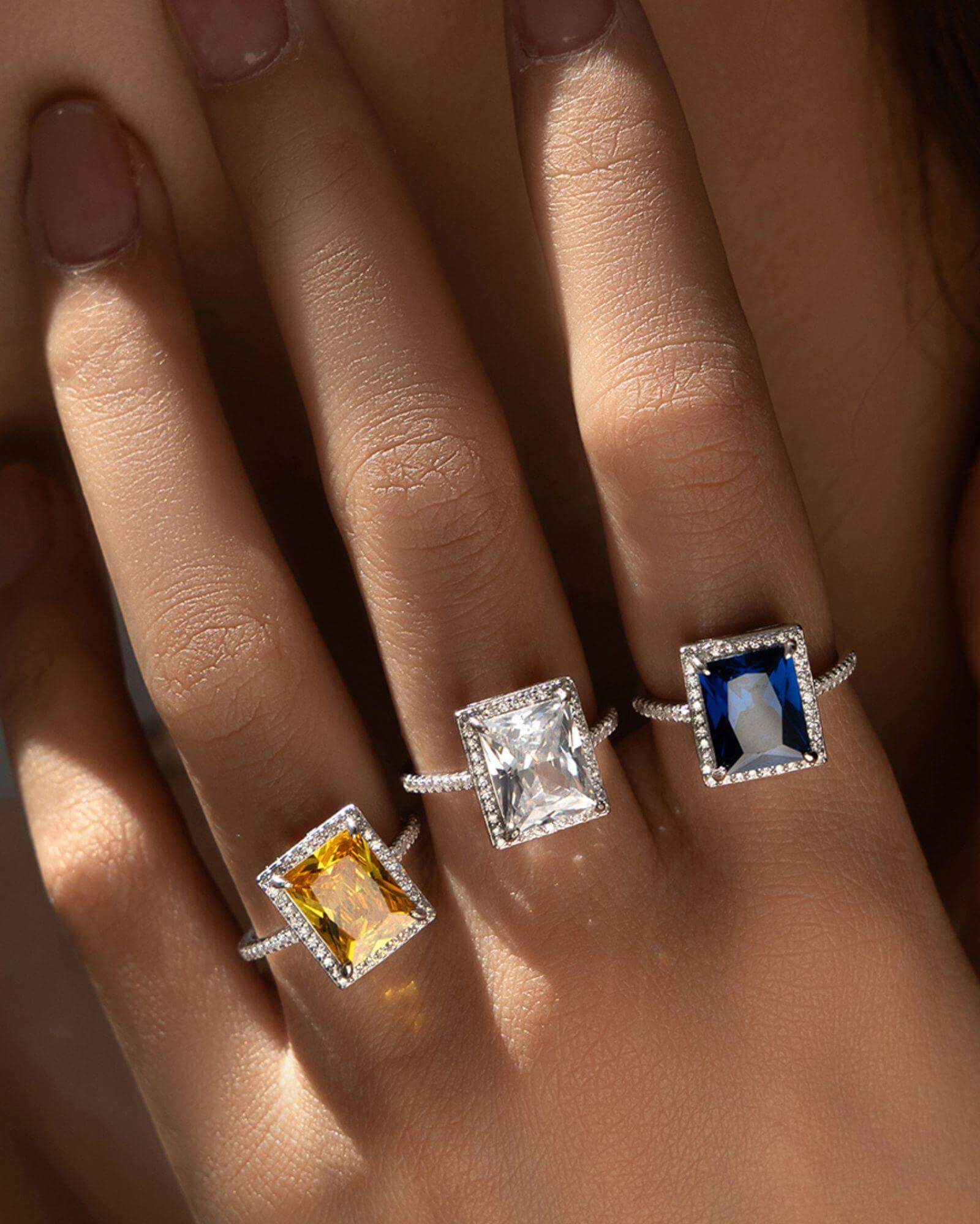 Three radiant cut rings featuring colorless, yellow, and blue stones set in sterling silver on a hand.