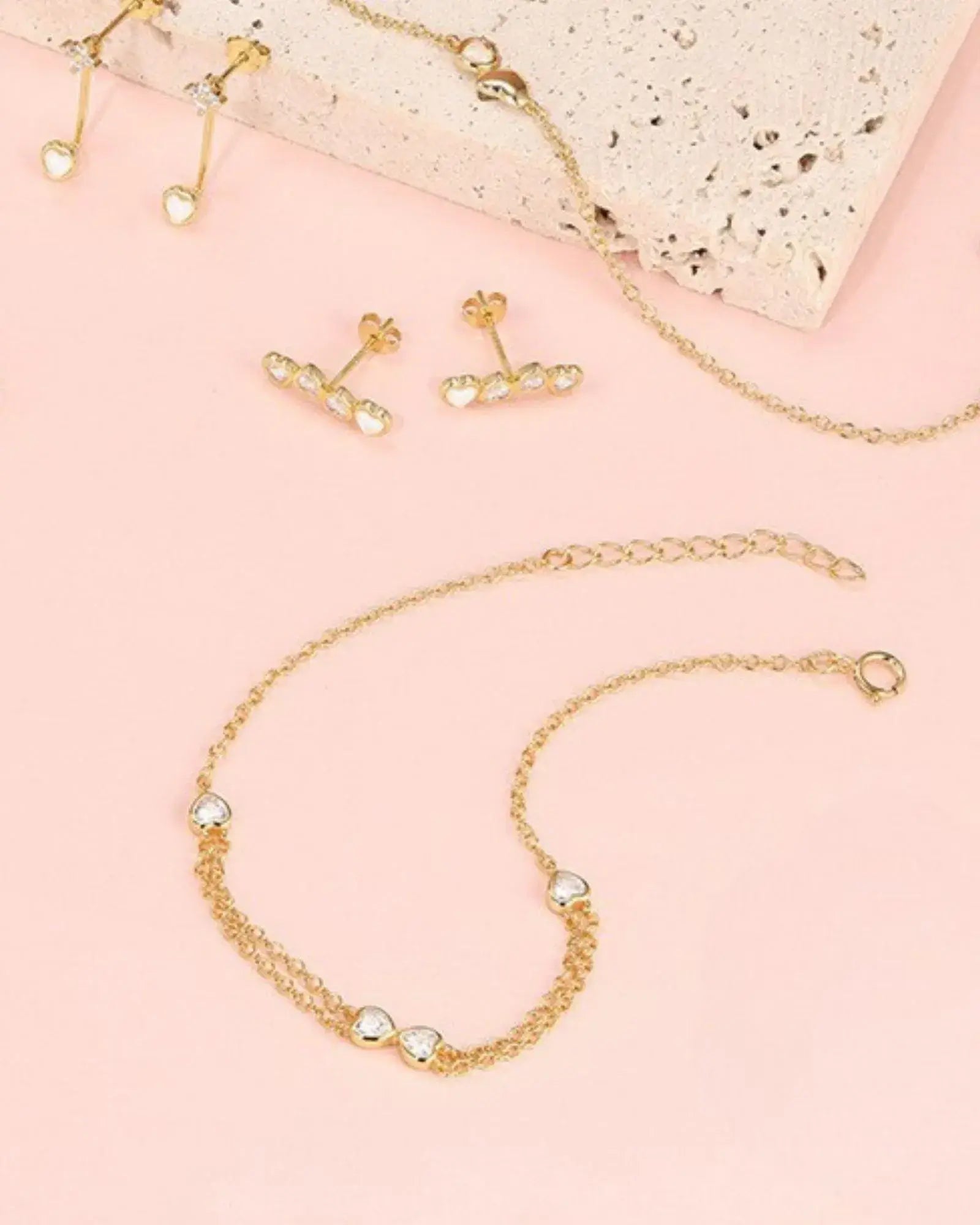 Elegant Heart Bow Bracelet with sparkling cubic zirconia centerpiece and matching gold earrings on a pink background.