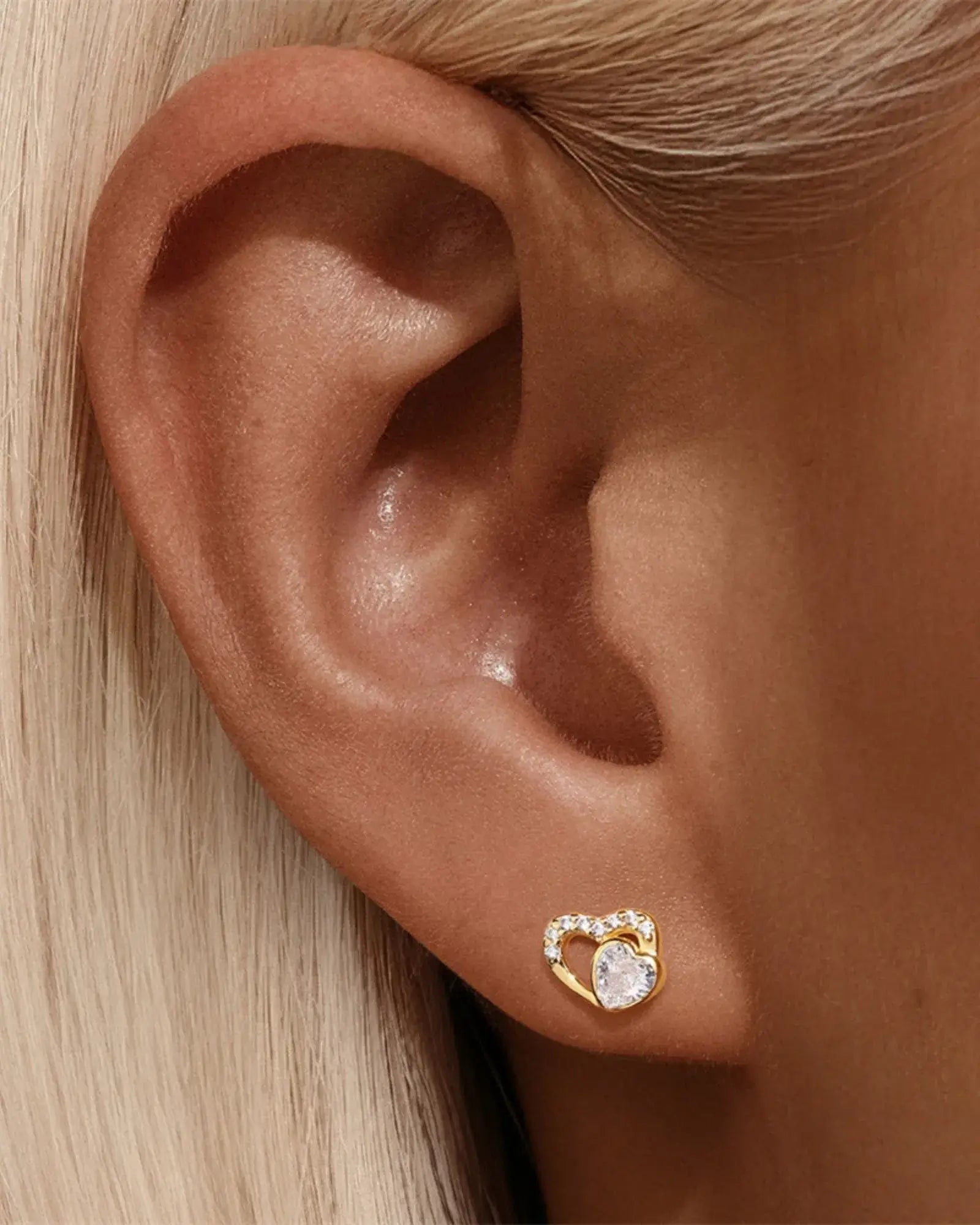 Close-up of a woman's ear wearing Love Glow Stud earrings in silver and gold with a heart design and sparkling stone.