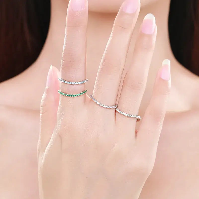 Close-up of a hand wearing multiple slim eternity bands, including silver and green zircon rings, showcasing elegant stacking style.