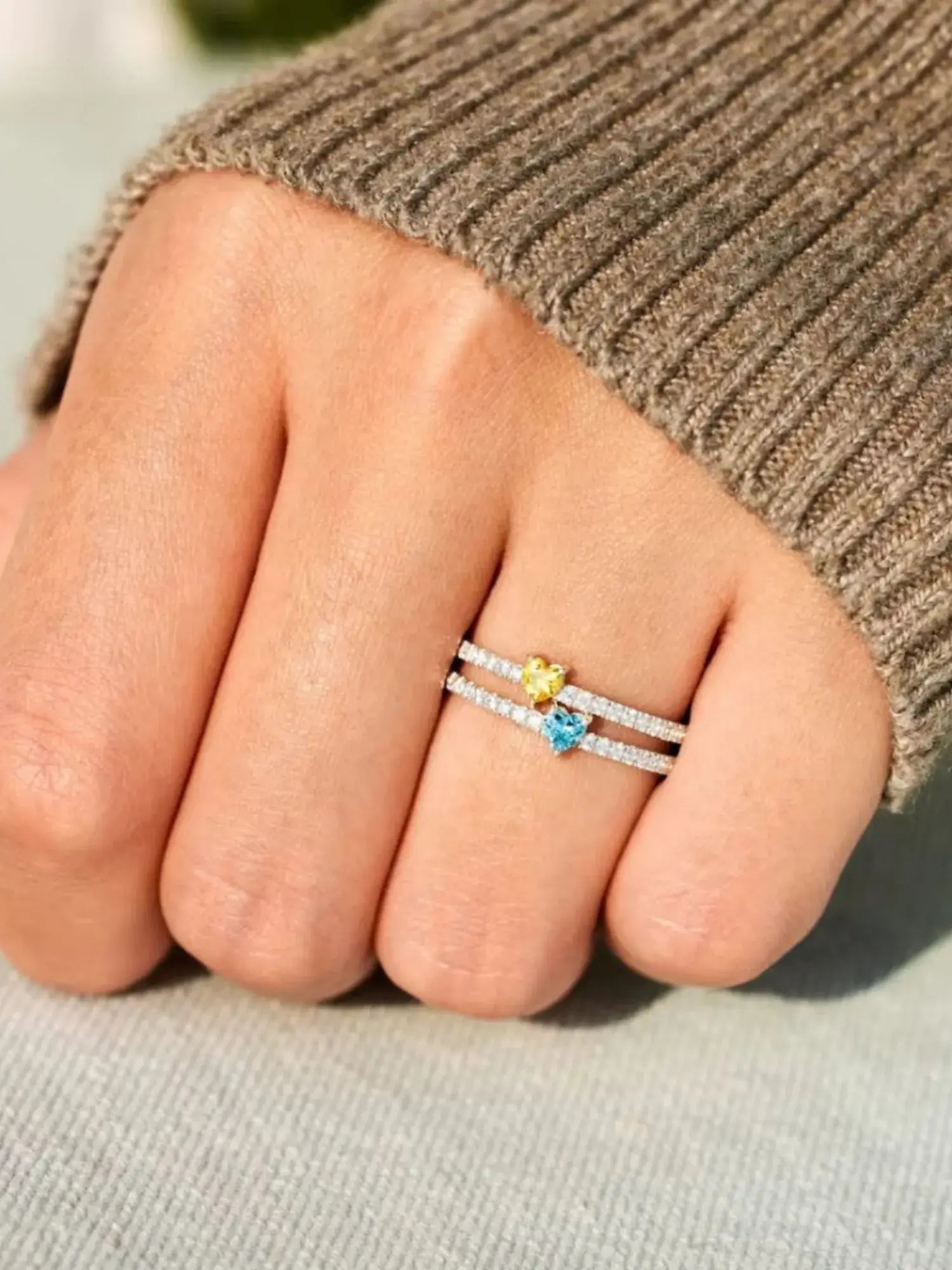 Close-up of a hand wearing a Two Heart Colourful Zircon Ring featuring yellow and blue zircon stones on a textured surface.