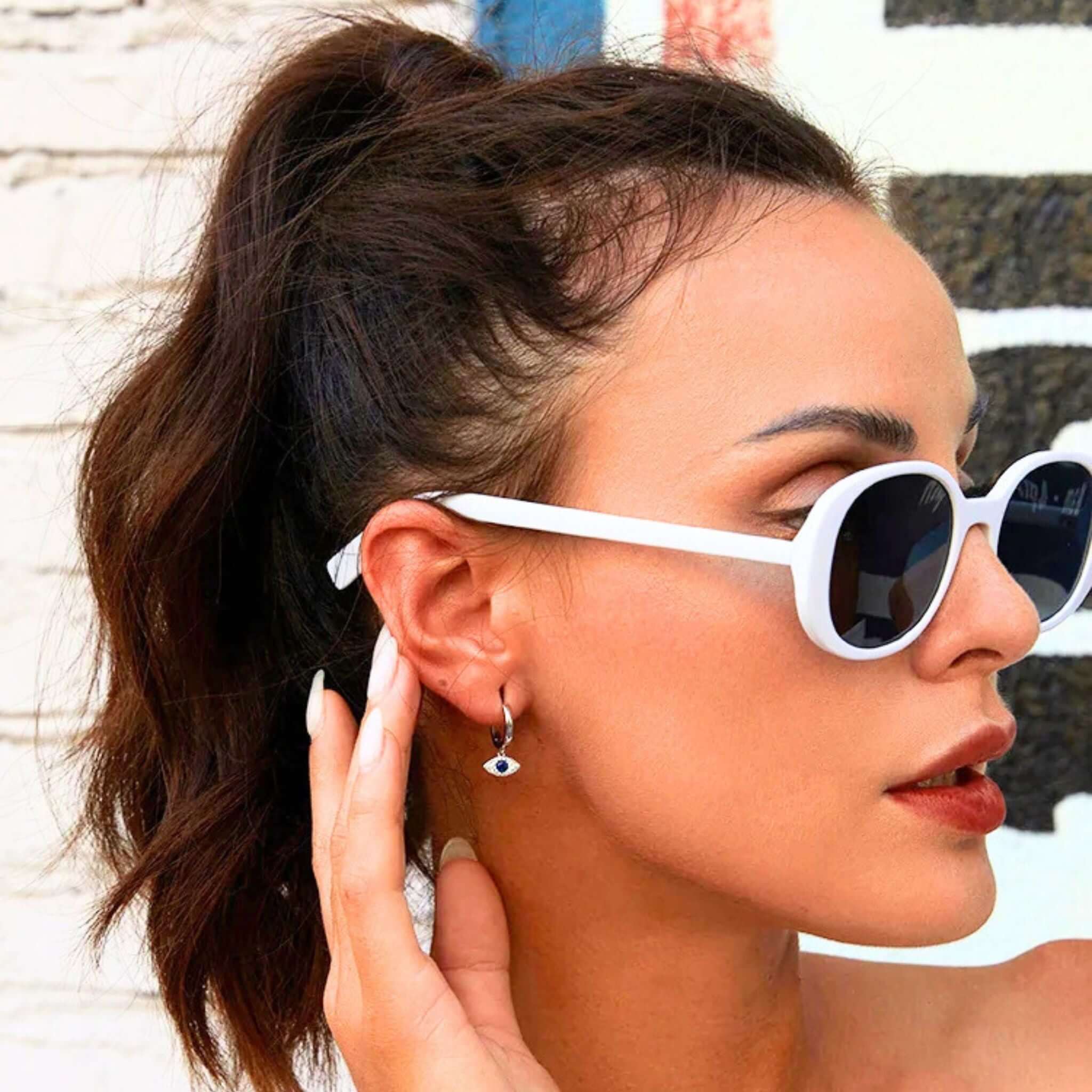 Woman wearing white sunglasses and blue eye earrings, showcasing a trendy summer look.