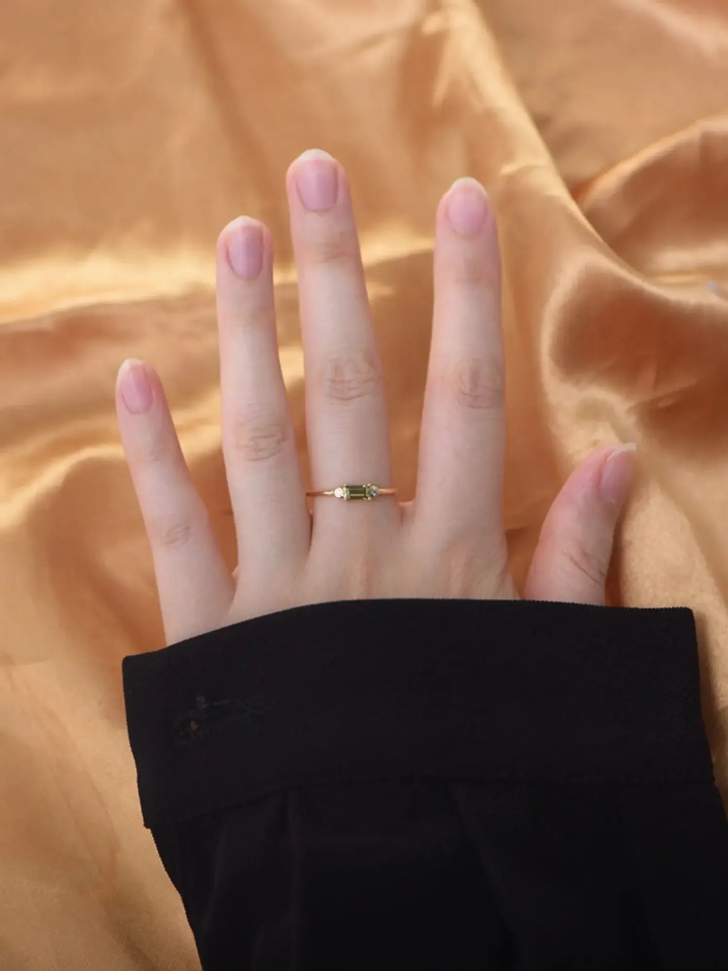 Hand displaying Viola Ring, featuring a green stone, set against a luxurious golden fabric backdrop.