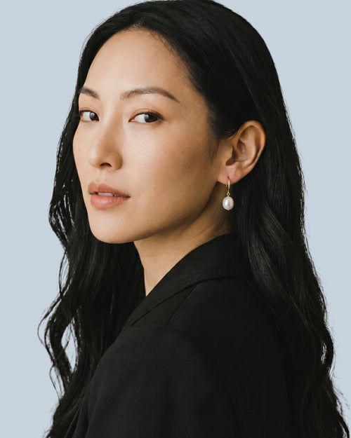 Woman with long dark hair wearing a black blazer and a white pearl drop earring against a light blue background