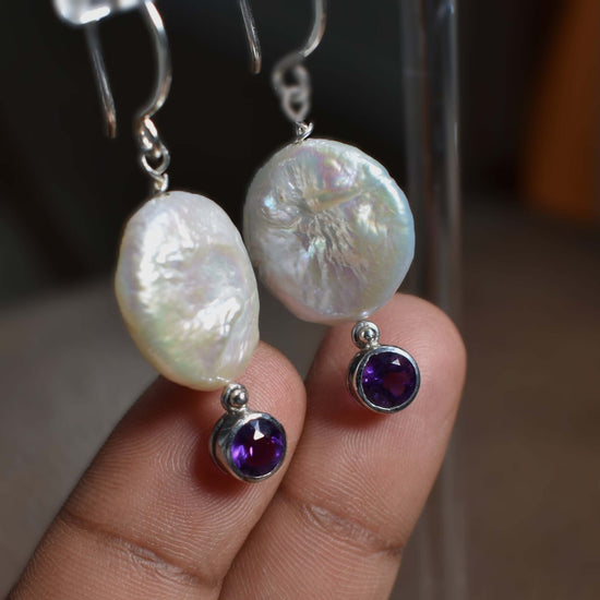 A pair of white freshwater coin pearls and Amethyst connector is shown against two fingers in order to compare the size 