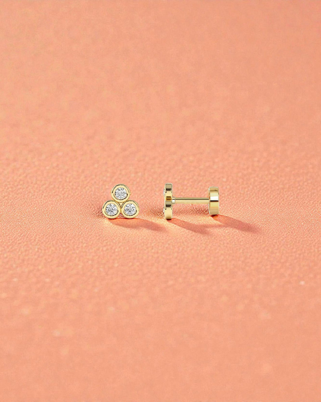 Pearlystones gold three stone cubic zirconia flat back stud earring for cartilage piercings on a peach background.