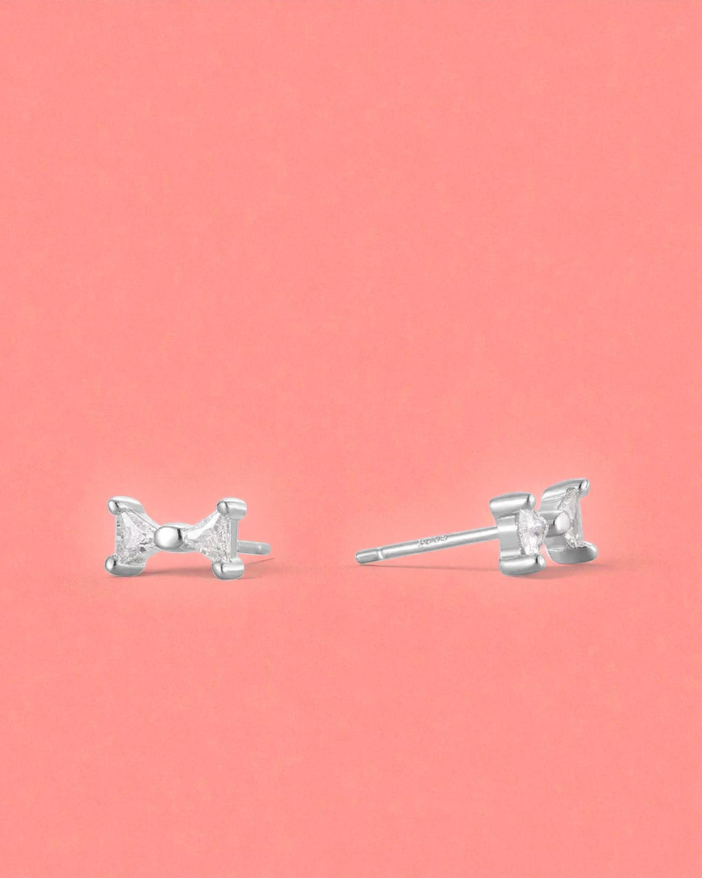 Small Bow Earrings in Sterling Silver studs on a pink background, offering elegant and lightweight style.