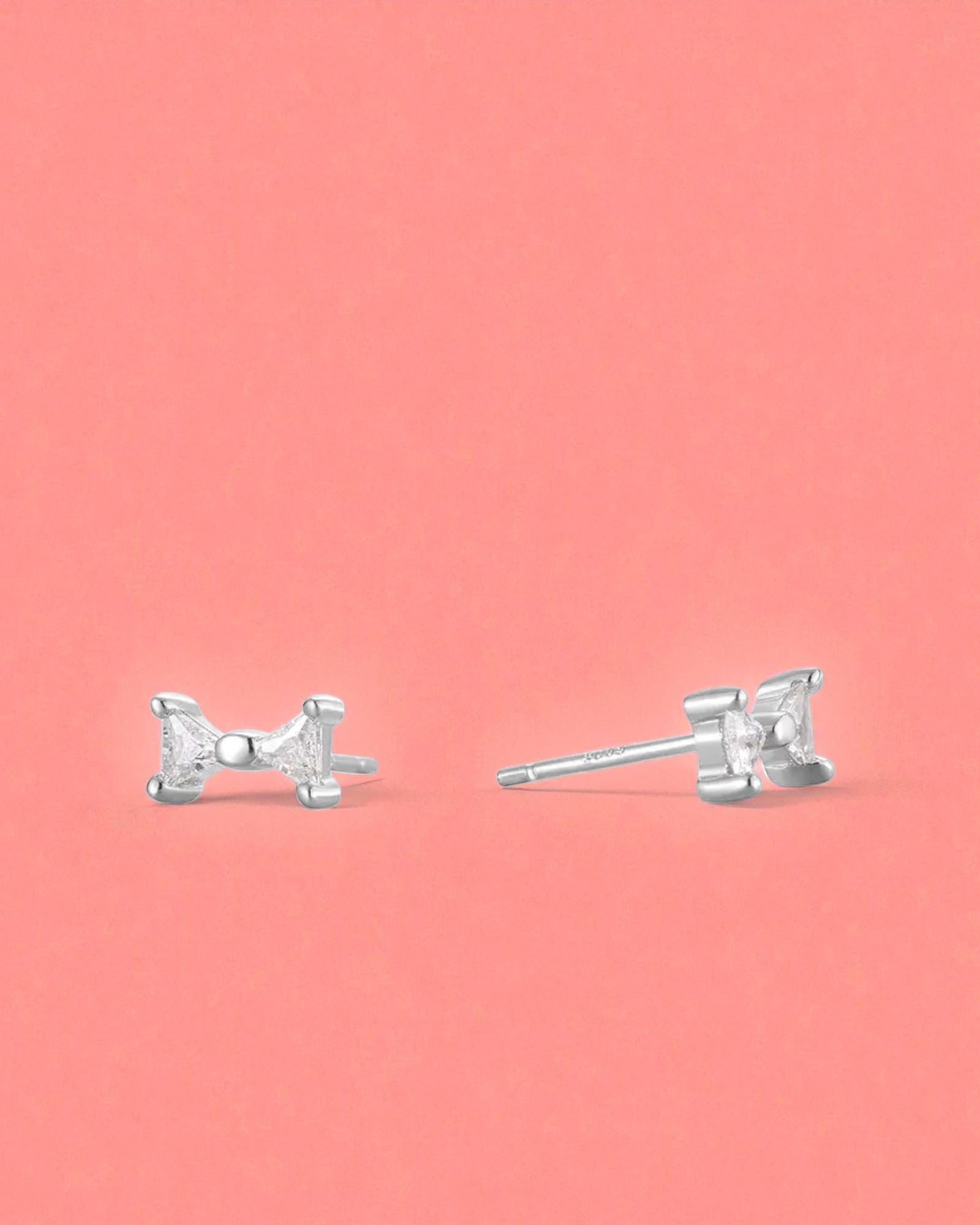 Small Bow Earrings in Sterling Silver studs on a pink background, offering elegant and lightweight style.