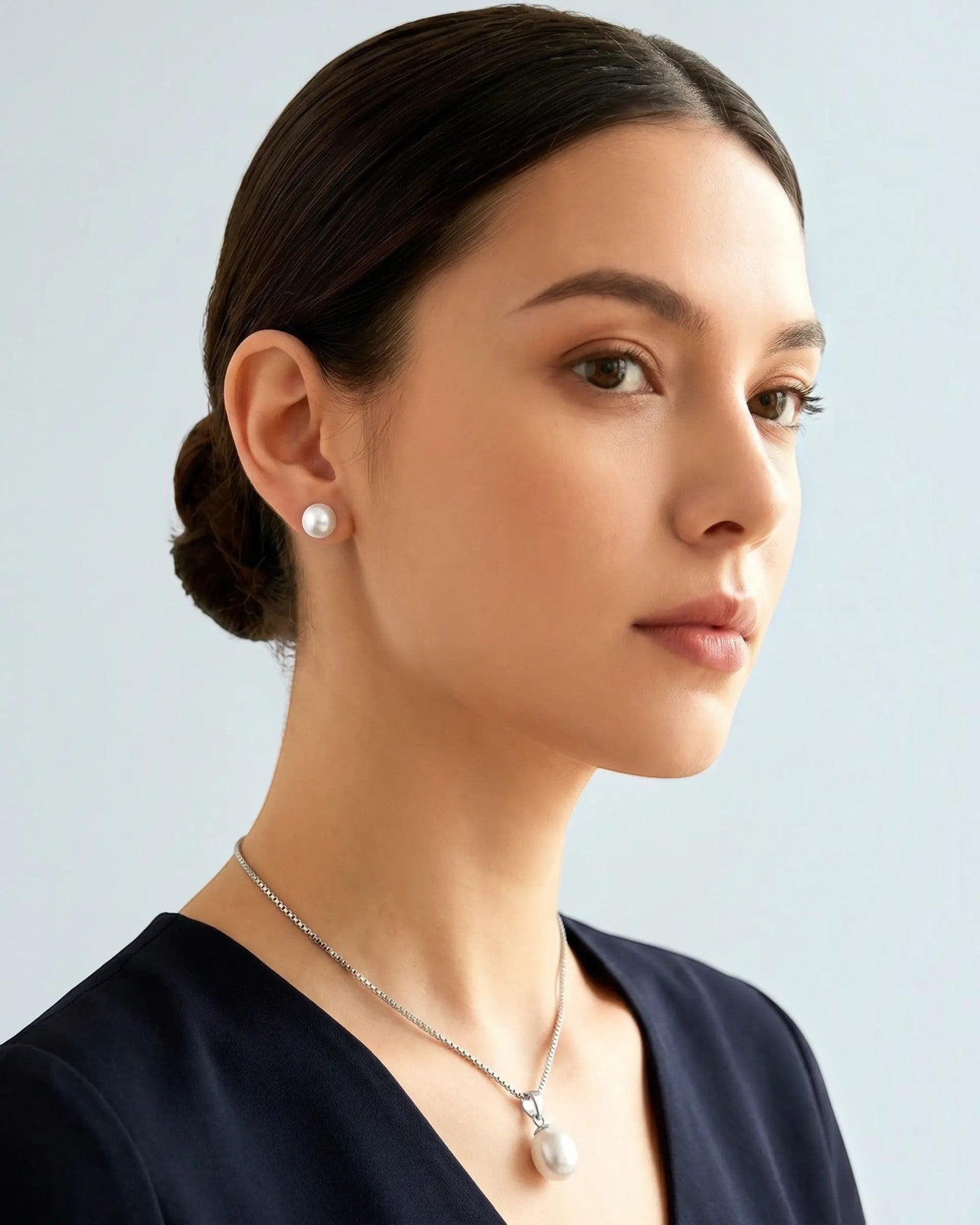A woman with dark hair in a low bun is shown in profile wearing a silver chain necklace with a white pearl pendant and a matching pearl stud earring, against a light gray background.