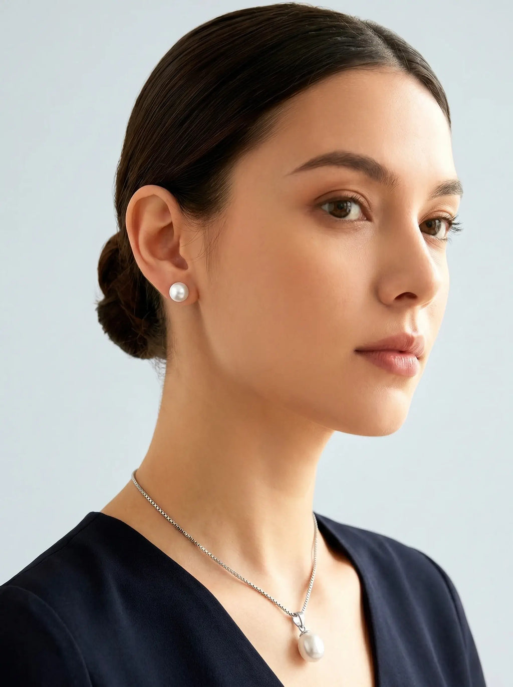 A woman with dark hair in a low bun is shown in profile wearing a silver chain necklace with a white pearl pendant and a matching pearl stud earring, against a light gray background.